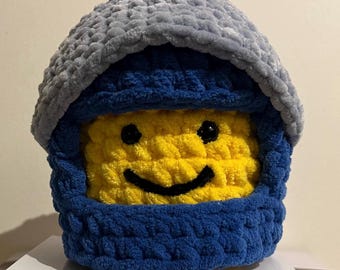 Puede incluir: Un gorro tejido a mano en forma de cabeza de figura Lego. El gorro presenta una cara amarilla con una sonrisa y ojos negros, enmarcada por una capucha azul y gris. La textura suave y afelpada es visible.