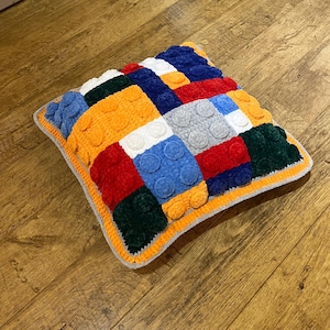 Könnte beinhalten: Ein quadratisches, handgefertigtes Kissen mit einem farbenfrohen Patchwork-Design. Das Kissen hat verschiedene Quadrate in Orange, Rot, Blau, Grün, Weiß und Grau mit erhabenen kreisförmigen Details. Das Kissen hat einen orangefarbenen Rand und eine graue Kante.