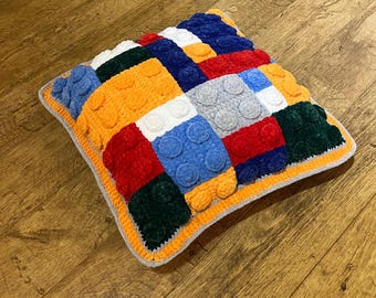 Puede incluir: Un cojín cuadrado hecho a mano con un diseño de patchwork colorido. El cojín presenta varios cuadrados de color naranja, rojo, azul, verde, blanco y gris, con detalles circulares en relieve. El cojín tiene un borde naranja y un borde gris.