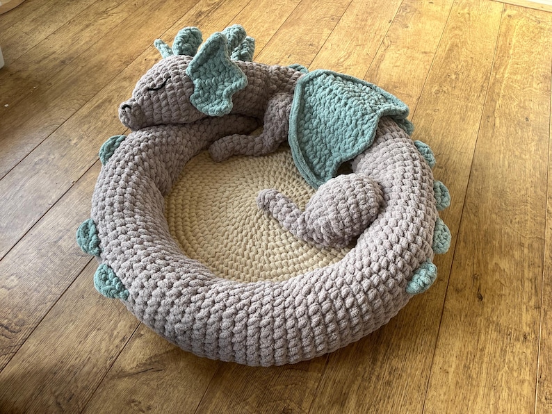 Puede incluir: Una cama para mascotas hecha a mano con ganchillo en forma de drag&oacute;n. La cama es gris con detalles turquesa en las alas y p&uacute;as. El centro es de color beige claro, proporcionando un espacio acogedor para una mascota peque&ntilde;a.