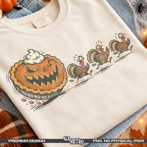 May include: A cream-colored t-shirt featuring a graphic illustration of a jack-o'-lantern pumpkin pie with whipped cream, being chased by three brown turkeys. The design includes motion lines and a distressed, vintage-style artistic aesthetic.