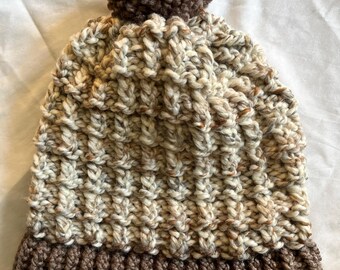 Può includere: Un berretto lavorato a mano a maglia con un motivo strutturato nei toni del beige, marrone e crema. Il cappello presenta un bordo marrone a coste e un pompon abbinato sulla parte superiore. Questo caldo cappello invernale è perfetto per il freddo.