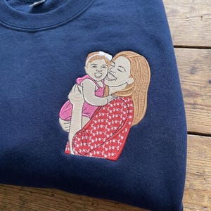 Puede incluir: Sudadera azul marino con un diseño bordado de una persona sosteniendo a un niño. El diseño presenta a una persona con cabello largo castaño claro que usa una blusa roja estampada, sosteniendo a un niño con un atuendo rosa. El diseño está cosido en tonos de rojo, rosa y marrón.