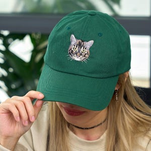 May include: A dark green baseball cap with an embroidered cat face on the front. The cat's face is detailed with brown and gray stripes, yellow eyes, and white whiskers. The cap has a curved brim and a button on top.