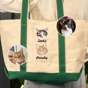 May include: A cream-colored tote bag with green trim and handles. The bag features embroidered cat portraits and the names "Sophie" and "Pawsley" below the images. Two additional cat portraits are in circular frames on the bag.