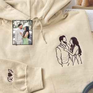 以下が含まれることがあります： 胸元にカップルのダークブラウンの刺繍、袖にハートマークとRとJのイニシャルが刺繍されたベージュのパーカー。このデザインは、挿入された小さなカップルの写真をもとに刺繍されています。