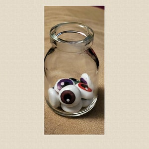 May include: A glass jar filled with white plastic eyeballs with colorful irises. The jar is on a brown surface.