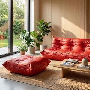 May include: A contemporary living room with a red leather sofa and footstool. The space is furnished with a wooden coffee table and potted plants. A large window provides a view of the garden, creating a bright and open feel.