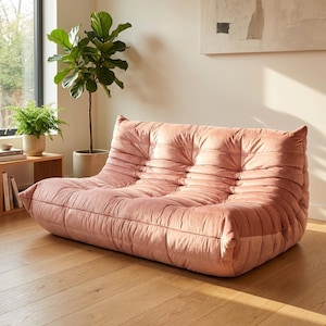 May include: A blush-pink sofa with a distinctive tufted design, set on a wooden floor. The room includes a large window, plants, and artwork. The sofa's low profile and soft texture create a comfortable aesthetic.
