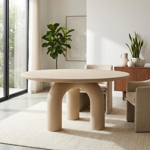 Puede incluir: Una mesa de comedor redonda beige con un diseño único de patas arqueadas. La mesa está sobre una alfombra color crema en una habitación bien iluminada. El diámetro de la mesa es de aproximadamente 122 cm.