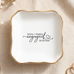 May include: A square white ceramic trinket dish with a scalloped edge and a gold-painted rim. The center features black text that reads Henry + Sophia engaged 06.20.2026 with a small heart graphic at the end of the word engaged.