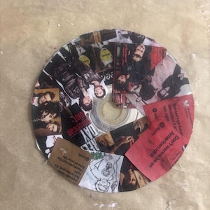 May include: A circular compact disc decorated with a collage of Green Day imagery and text. The surface features snippets of album art, band members, and lyrics including 'Don't wanna be an American idiot' and 'But in the end, it's right'.