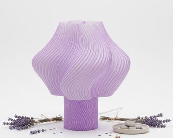 May include: A lavender-colored table lamp with a unique, wavy design. The lamp has a cylindrical base and a bulbous shade. The image also includes sprigs of lavender and a small dish with stones.