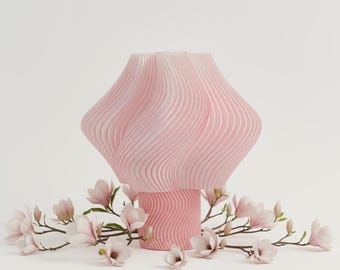 May include: A pink, wavy-textured vase with a flared top and a cylindrical base. The vase is surrounded by magnolia branches with pink and white flowers, creating a soft, floral arrangement. The vase is a decorative home decor item.