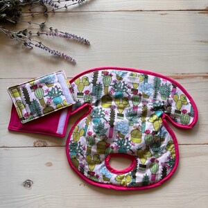 May include: A baby car seat head support and matching small wallet, both with a cactus print on a light pink background. The head support has a bright pink trim. The wallet has a velcro closure.