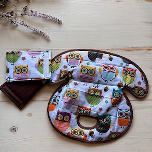 May include: A collection of owl-patterned accessories, possibly a wallet and a curved item, perhaps a heating pad. The items display a light blue backdrop adorned with vibrant owl illustrations in shades of orange, green, and purple. Brown edging frames the pieces.
