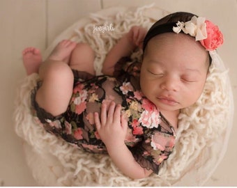 May include: A newborn baby girl sleeping in a white bowl with a floral lace onesie and a black headband with a pink flower.