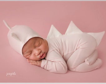 May include: A pink baby onesie with a dinosaur tail and matching hat. The baby is sleeping on a pink background.