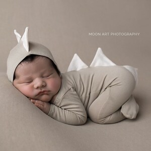 May include: A newborn baby wearing a beige dinosaur costume with white accents, sleeping on a beige background. The baby is wearing a hat with white dinosaur ears.