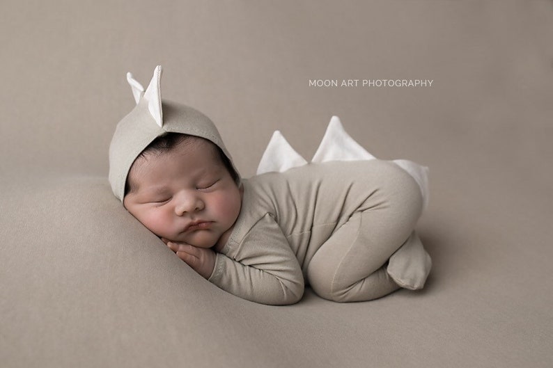 May include: A newborn baby wearing a beige dinosaur costume with white accents, sleeping on a beige background. The baby is wearing a hat with white dinosaur ears.
