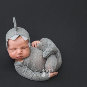 May include: A newborn baby wearing a gray shark costume with a white fin on the head, sleeping on a black background.