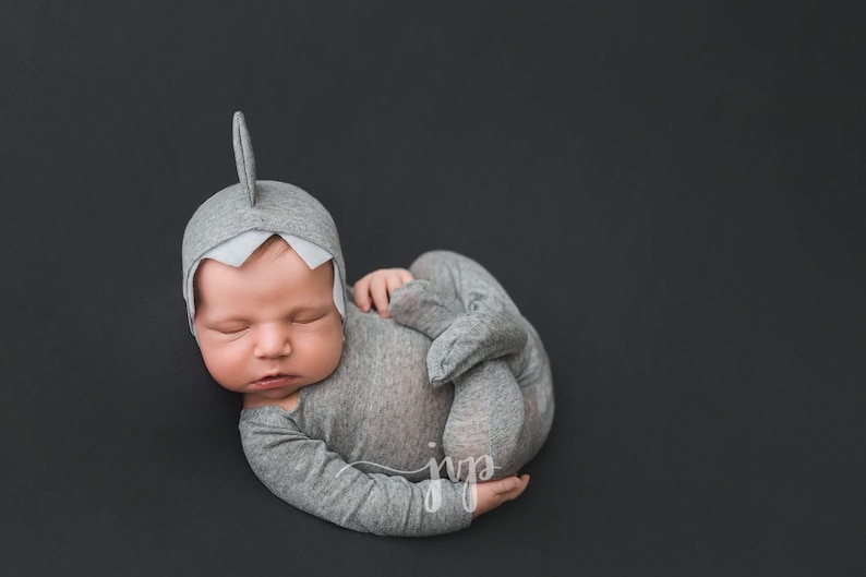 May include: A newborn baby wearing a gray shark costume with a white fin on the head, sleeping on a black background.