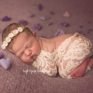 Puede incluir: Una recién nacida duerme sobre una manta morada con una envoltura de encaje blanco y una corona de flores blancas. Hay pequeños corazones de fieltro morado esparcidos alrededor del bebé.