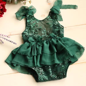 Op de afbeelding: Een donkergroene babyromper met een gelaagde rok en bloemenkanten details. De romper heeft delicate strikken op de schouders en taille. Rode bloemen en paarse takjes staan op de achtergrond.