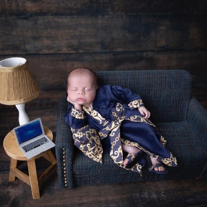 Op de afbeelding: Een slapende baby in een blauwe en gouden robe zit op een blauwe fluwelen bank. Een kleine houten tafel met een laptop en een lamp staat naast de bank.