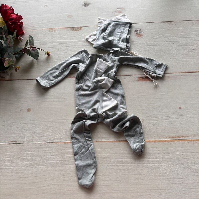 May include: A gray baby onesie with a white trim and a matching gray hat. The onesie has long sleeves and legs, and the hat has a pointed top.