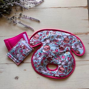 May include: A set of baby accessories featuring a whimsical pattern of unicorns, pugs, rainbows, and food items like burgers and ice cream on a light blue background. The set includes a neck pillow and a smaller square item, both with a pink trim.