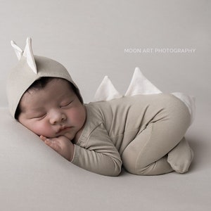 May include: A newborn baby wearing a beige dinosaur onesie with a white dinosaur tail and a beige hat with white dinosaur ears. The baby is sleeping on a white surface. MOON ART PHOTOGRAPHY is printed in the top right corner of the image.