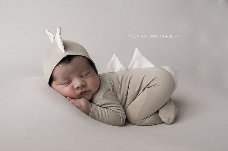 May include: A newborn baby wearing a beige dinosaur onesie with a white dinosaur tail and a beige hat with white dinosaur ears. The baby is sleeping on a white surface. MOON ART PHOTOGRAPHY is printed in the top right corner of the image.