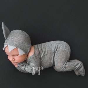 May include: A newborn baby wearing a gray shark costume with a white fin and teeth. The baby is lying on its side with its head resting on its arm.