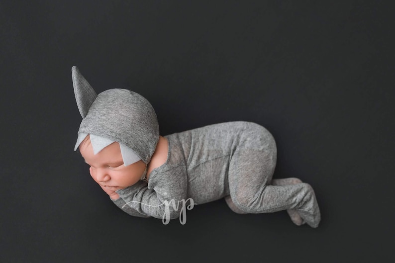 May include: A newborn baby wearing a gray shark costume with a white fin and teeth. The baby is lying on its side with its head resting on its arm.