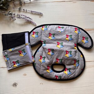 Puede incluir: Un conjunto de tres artículos de tela gris con un patrón del escudo de Hogwarts. El conjunto incluye una pieza grande y acolchada con un borde negro, una pieza rectangular pequeña y una pieza rectangular negra. El escudo de Hogwarts presenta los colores rojo, amarillo, verde y azul.