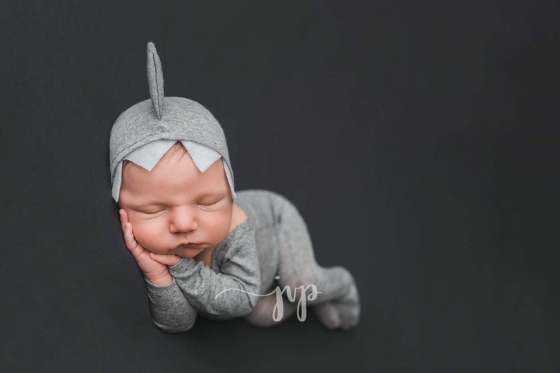 May include: A newborn baby wearing a gray shark costume is sleeping on a gray background. The baby is lying on its stomach with its head turned to the side. The baby's eyes are closed and its mouth is slightly open.