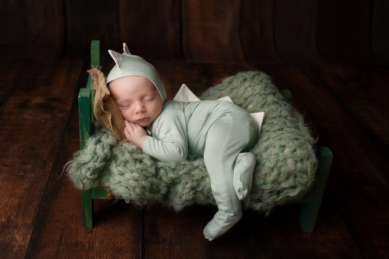 May include: A baby wearing a light green dinosaur costume sleeps on a green knitted blanket in a miniature wooden bed.