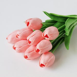 Puede incluir: Un ramo de tulipanes rosas con hojas verdes. Los tulipanes est&aacute;n en varias etapas de floraci&oacute;n, algunos cerrados y otros parcialmente abiertos, revelando un toque de rojo en el interior. El arreglo est&aacute; sobre un fondo blanco.
