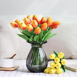 Puede incluir: Un ramo de tulipanes naranjas y amarillos en un jarr&oacute;n de cristal gris oscuro. Una taza de t&eacute; blanca con una cuchara de madera descansa sobre un posavasos de madera. Tambi&eacute;n hay tulipanes amarillos adicionales y un libro.