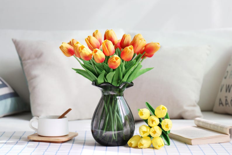 Puede incluir: Un ramo de tulipanes naranjas y amarillos en un jarr&oacute;n de cristal gris oscuro. Una taza de t&eacute; blanca con una cuchara de madera descansa sobre un posavasos de madera. Tambi&eacute;n hay tulipanes amarillos adicionales y un libro.