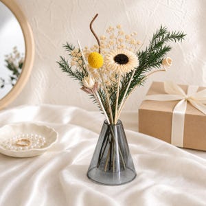 May include: A small, conical, translucent gray glass vase holds a bouquet of dried flowers, including a yellow billy ball, a cream-colored sunflower with a dark center, sprigs of baby's breath, green pine-like foliage, and a curled wooden stem.