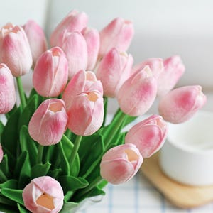 Puede incluir: Un ramo de tulipanes rosas con hojas verdes. Las flores est&aacute;n en plena floraci&oacute;n, con p&eacute;talos rosas suaves y centros amarillos. Una taza blanca y un posavasos de madera est&aacute;n en el fondo.