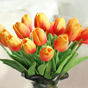 Puede incluir: Un ramo de tulipanes naranjas y amarillos con hojas verdes en un jarr&oacute;n de cristal oscuro. Las flores tienen un degradado de colores, pasando del amarillo al naranja y al rojo en las puntas.