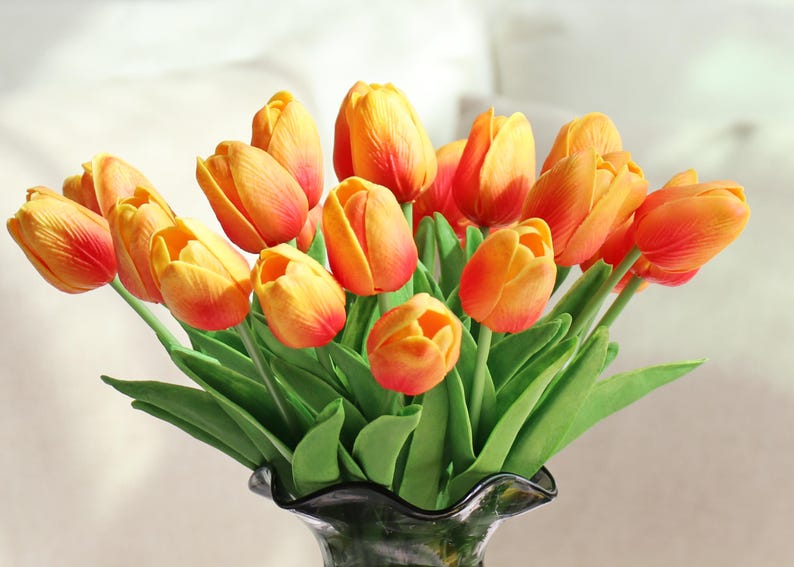Puede incluir: Un ramo de tulipanes naranjas y amarillos con hojas verdes en un jarr&oacute;n de cristal oscuro. Las flores tienen un degradado de colores, pasando del amarillo al naranja y al rojo en las puntas.