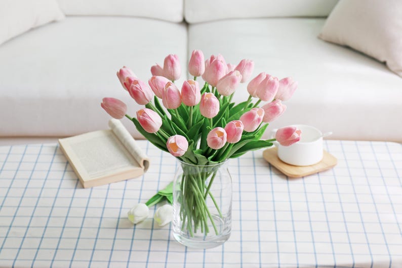 Puede incluir: Un ramo de tulipanes rosas en un jarr&oacute;n de cristal transparente. Los tulipanes tienen tallos y hojas verdes. El jarr&oacute;n est&aacute; sobre una mesa con un mantel a cuadros azules y blancos. Tambi&eacute;n hay un libro y un cuenco blanco.