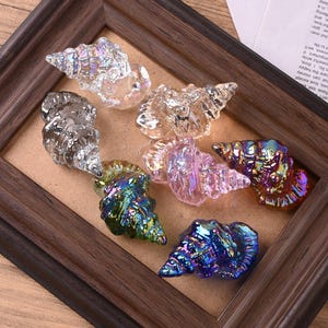 May include: A collection of iridescent glass seashell figurines in various colors, including clear, silver, pink, blue, and green. The seashells are displayed inside a dark brown wooden picture frame, creating a decorative display.