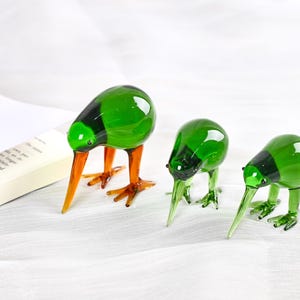 May include: Three glass kiwi bird ornaments. The birds have green bodies with black accents, orange beaks and legs. The figurines are on a white surface, with a book in the background.