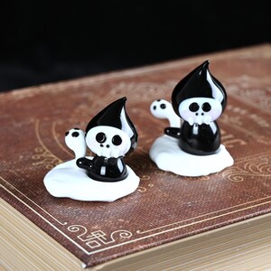 以下が含まれることがあります： 黒と白の小さな死神のようなキャラクターのフィギュアが2つ。各フィギュアは、黒い尖ったフード、骸骨の顔、黒い体を持っています。白い雲のようなベースに座っています。フィギュアは茶色のアンティーク調の本の上にあります。