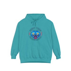 May include: A turquoise hoodie with a front pocket and drawstring hood. The hoodie features a circular graphic with a cartoon tiger face and the words "DERPY TIGER DELIVERY SERVICE". The tiger has blue eyes and a pink tongue.